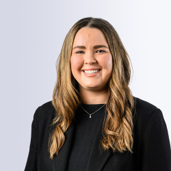 A young woman with long, wavy hair smiles brightly, wearing a black blazer over a dark top. She is positioned against a light gray background, exuding a professional and approachable demeanor.