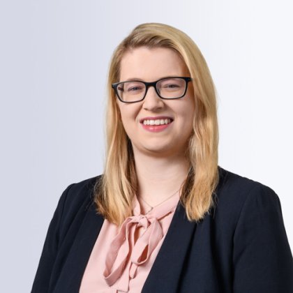 Portrait of a smiling woman with straight blonde hair, wearing black glasses, a light pink blouse with a bow tie, and a dark blazer, set against a light gray background.