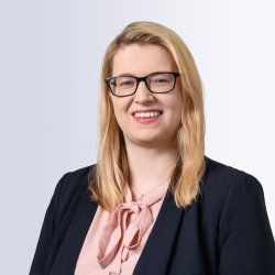 Portrait of a smiling woman with straight blonde hair, wearing black glasses, a light pink blouse with a bow tie, and a dark blazer, set against a light gray background.