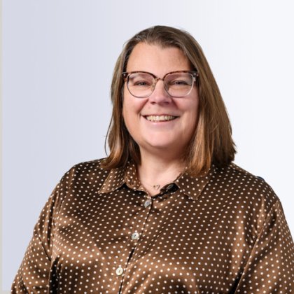 Portrait of a smiling woman with shoulder-length brown hair, wearing glasses and a brown satin blouse with white polka dots, against a light gray background.